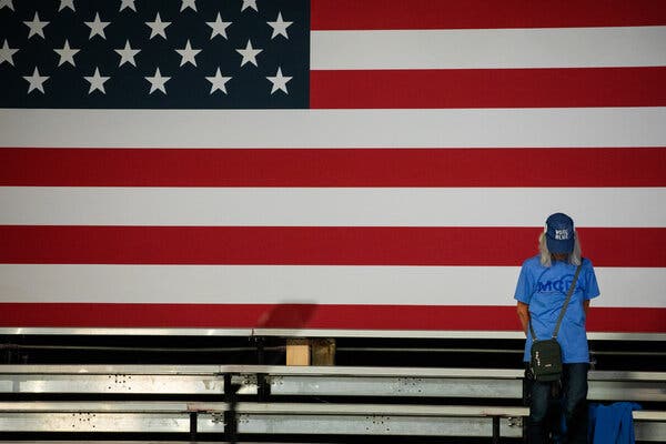 A person wearing a “Vote Blue” baseball cap stands in a row of bleachers in front of an enormous American flag.
